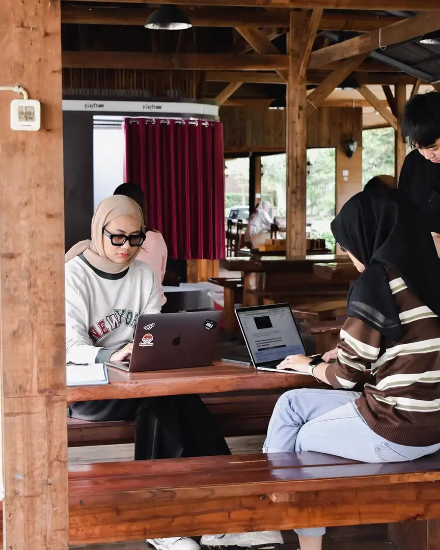 Tamu bekerja dengan laptop di meja kayu panjang di area saung