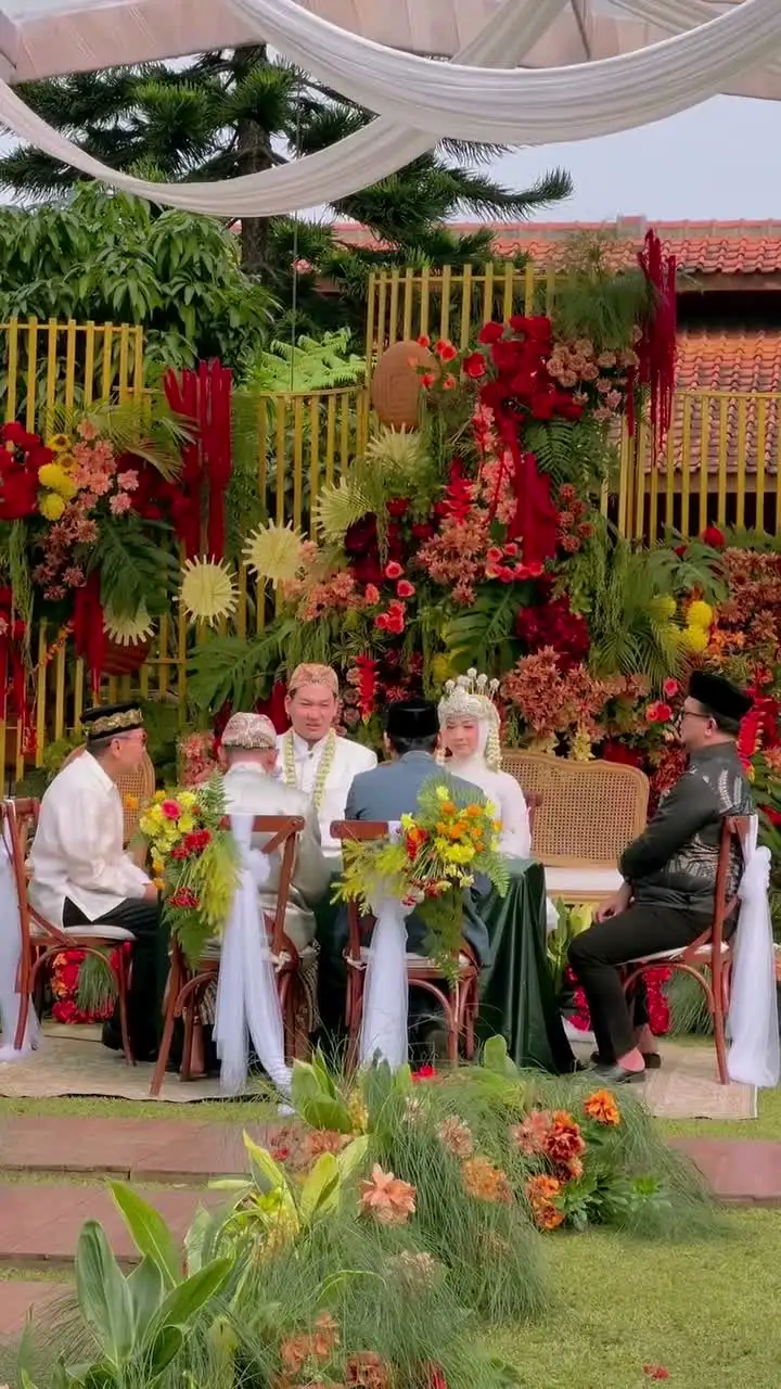 Pasangan pengantin berjalan di rumput di bawah payung kehormatan emas disaksikan para tamu