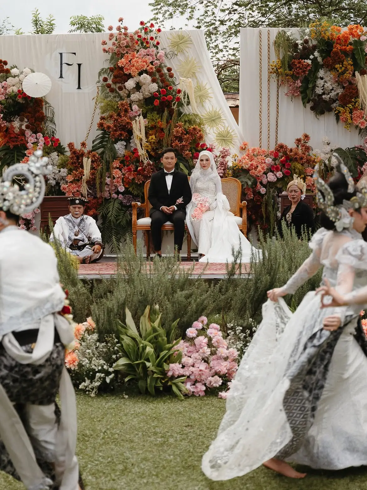 Pelaminan berhias bunga di atas rumput dengan pengantin duduk dan penari tampil